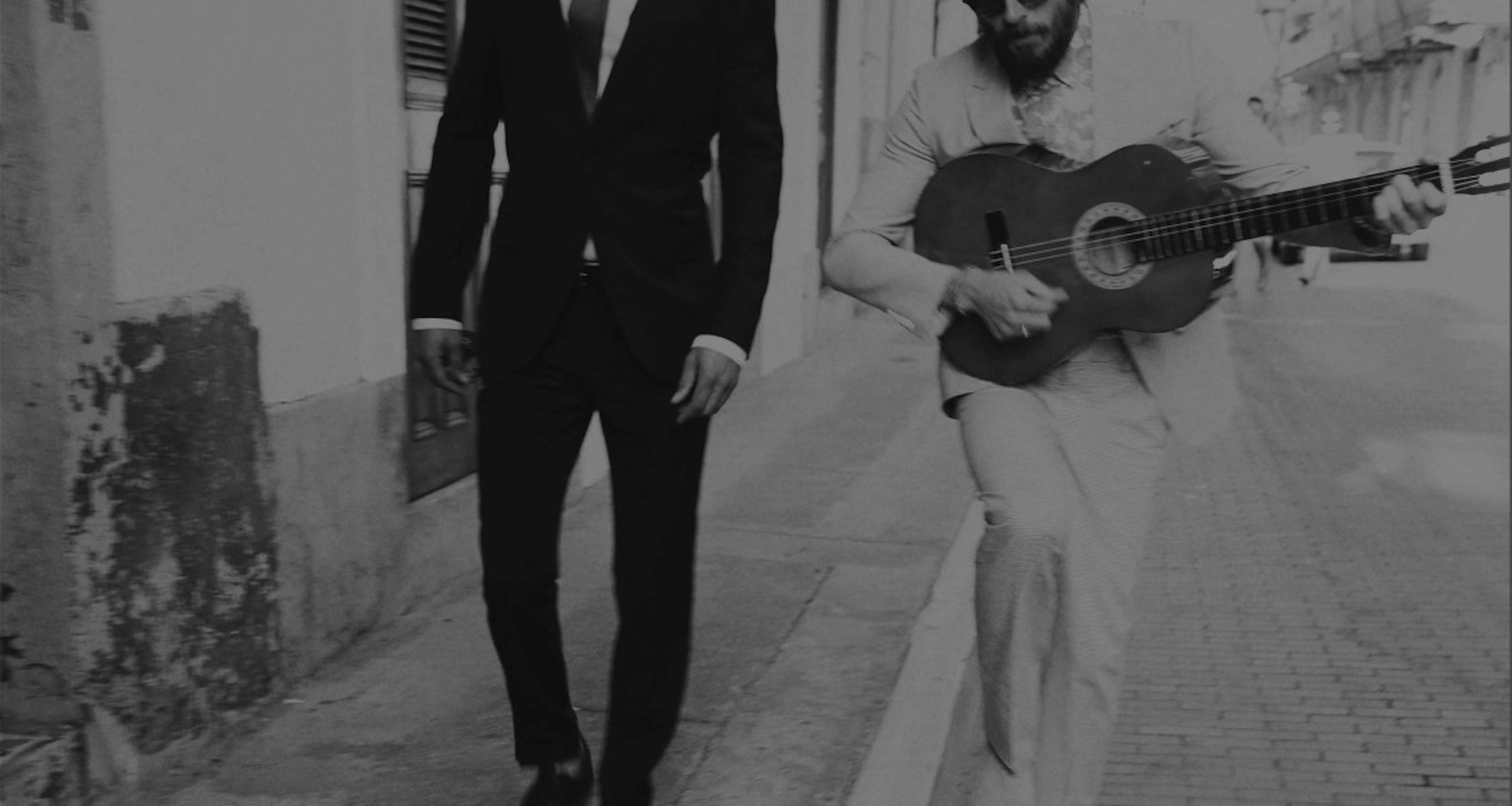 Image of 2 men in suits walking down the block, man on the right has a guitar.